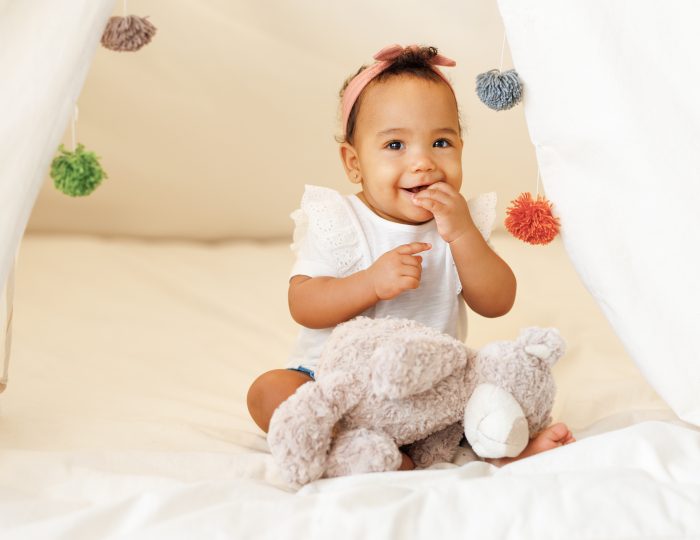 Portrait of a young baby girl smiling with a teddy bear sat on a bed.