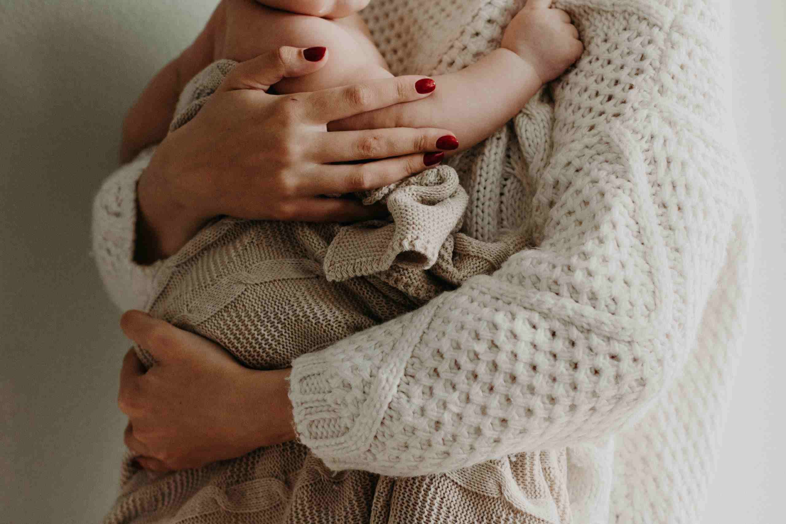 A mother with red nails holding her newborn baby in biege clothing.