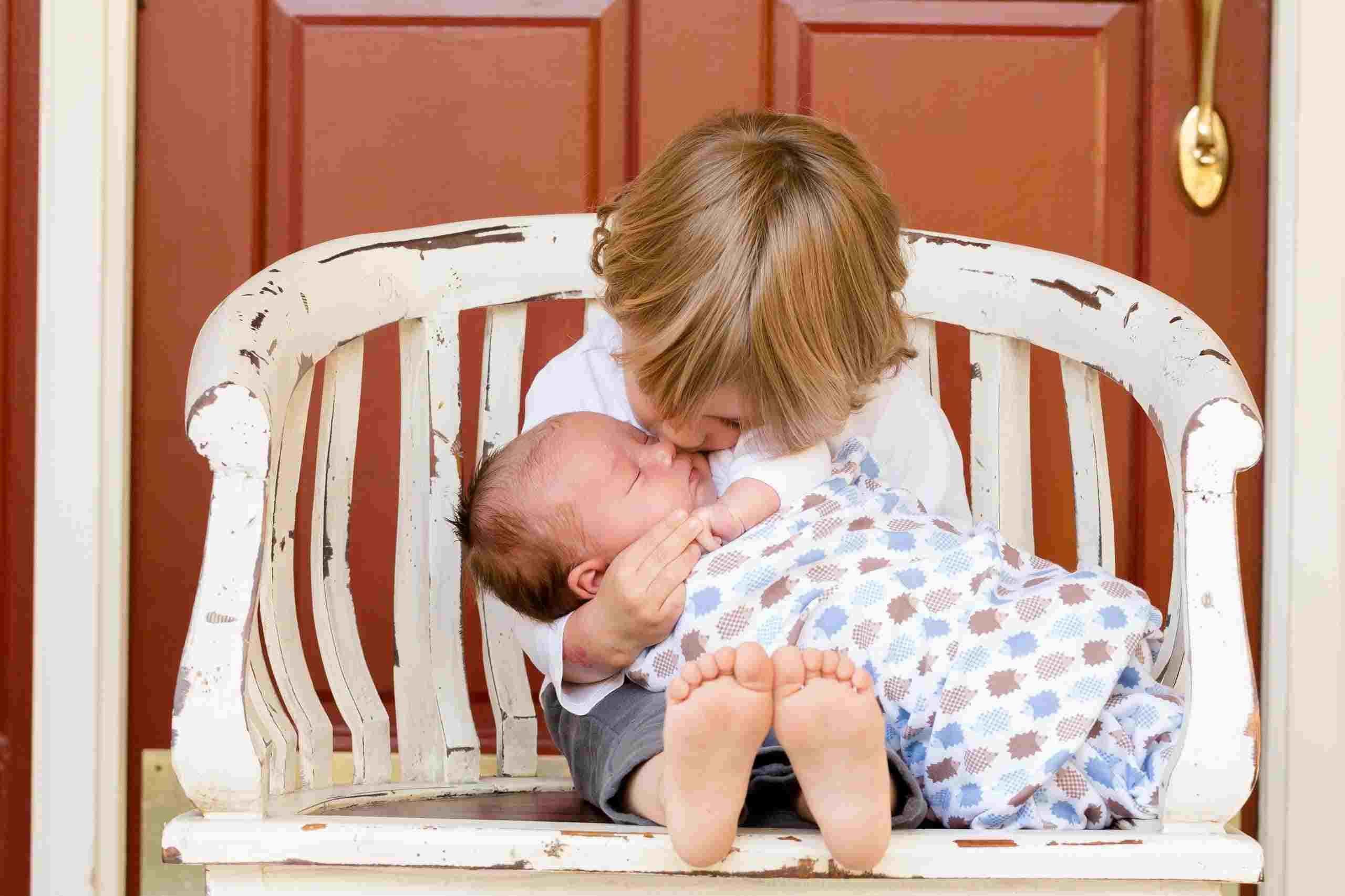 A toddler sat on a chair holding his baby sibling and kissing on the cheek.